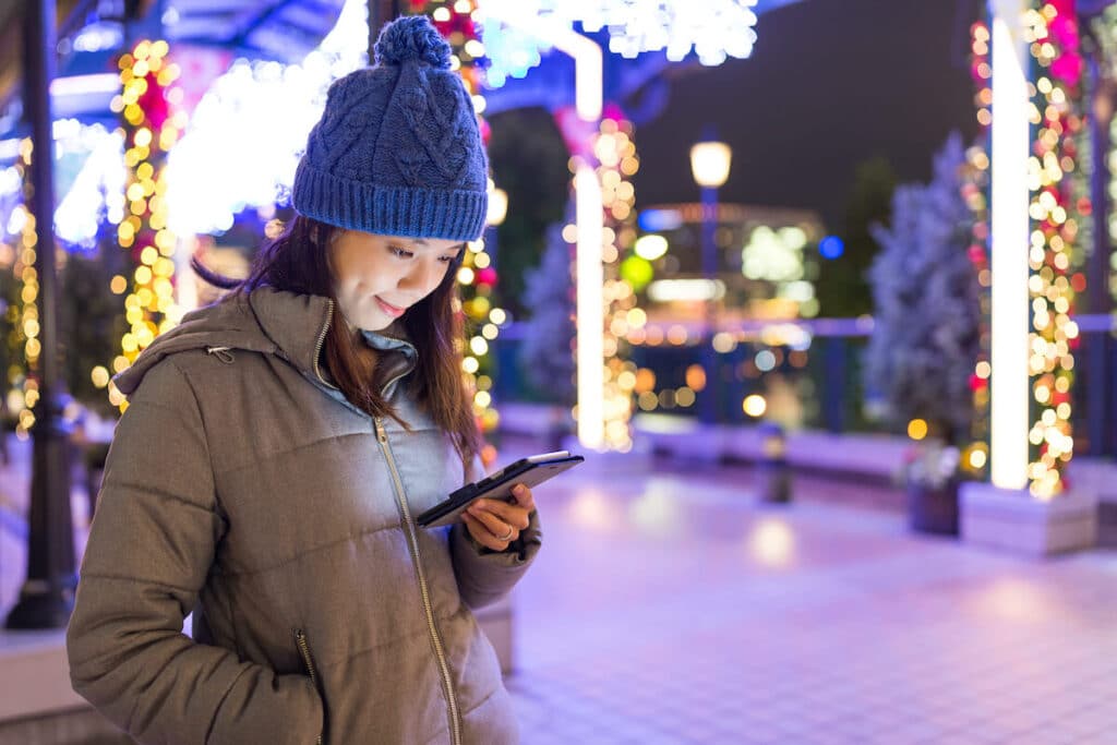 A woman in a hat and coat looks at her cellphone while she stands outside with Christmas lights in the background