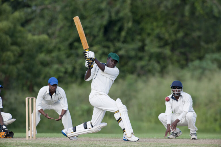 "Cricket action, batsman playing a shot, watched by fielders and the wicketkeeper."