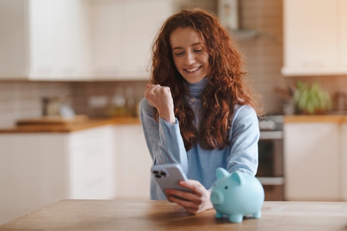 Happy Woman Checking Phone In Kitchen With Piggy Bank