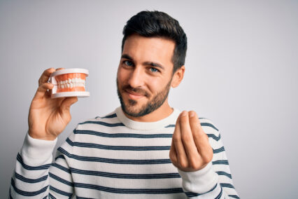 Young man with beard holding plastic denture teeth over white background doing money gesture with hands.