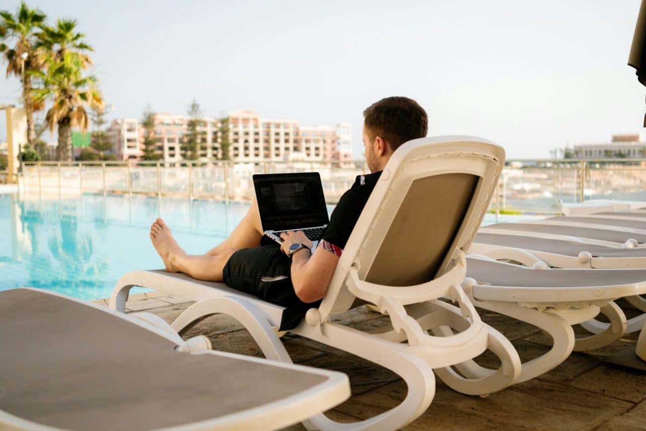 Person using a laptop on a sun lounger beside a pool.