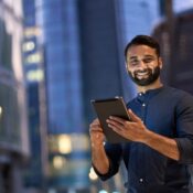 Smiling happy young business man professional manager standing outdoor on street holding using digital tablet online fintech in night city with urban lights