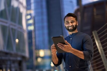 Smiling happy young business man professional manager standing outdoor on street holding using digital tablet online fintech in night city with urban lights