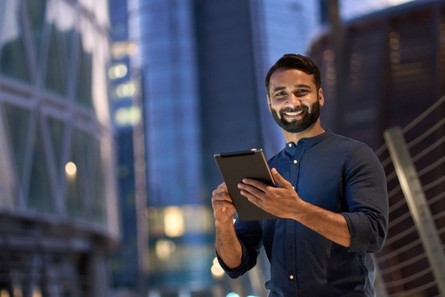 Smiling happy young business man professional manager standing outdoor on street holding using digital tablet online fintech in night city with urban lights