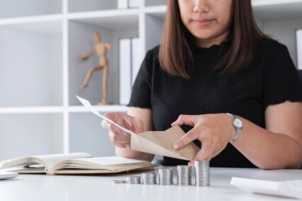 Young Woman Planning to Save Money by Counting Coins and Organizing Finances at Home Office Desk with Notebook and Envelopes