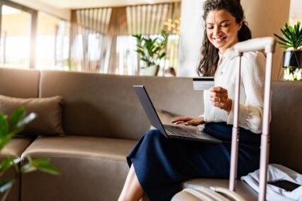 A woman sits on a sofa in a modern lobby with her laptop and luggage. She smiles while holding a credit card, ready to make an online purchase or booking.