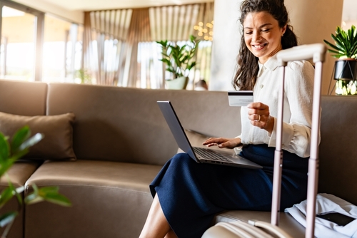 A woman sits on a sofa in a modern lobby with her laptop and luggage. She smiles while holding a credit card, ready to make an online purchase or booking.
