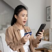 A woman staring at cellphone screen with shocked expression while holding payment card
