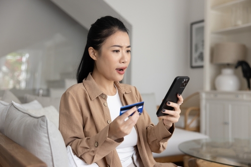 A woman staring at cellphone screen with shocked expression while holding payment card