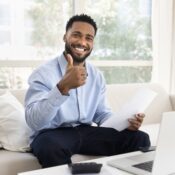 Man sitting on sofa at home, looking at camera with joyful expression, holding legal paper document feeling satisfied with receiving news