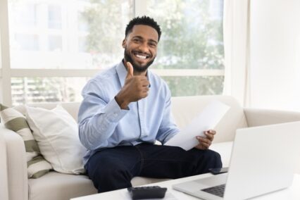 Man sitting on sofa at home, looking at camera with joyful expression, holding legal paper document feeling satisfied with receiving news