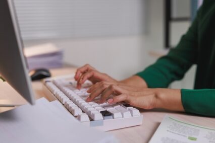 A person's hands are actively typing on a modern white keyboard while working at a desk