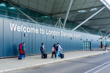 Welcome to London Stansted Airport sign with a group of travellers walking towards departures.