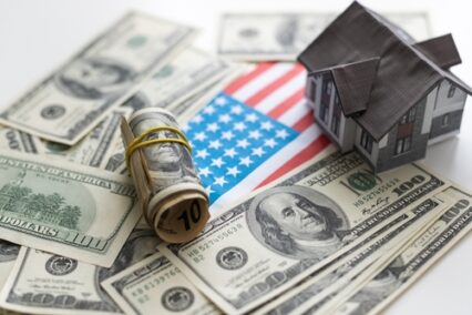 A toy house made of wood with a roof costs money, on a hundred-dollar bill.