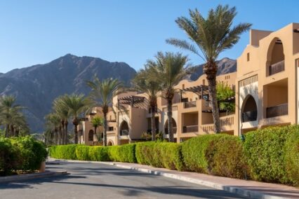 Row of Arabic style modern townhouses, residential architecture with green vegetation and palm trees around, Hajar mountains in the background, Fujairah, United Arab Emirates.