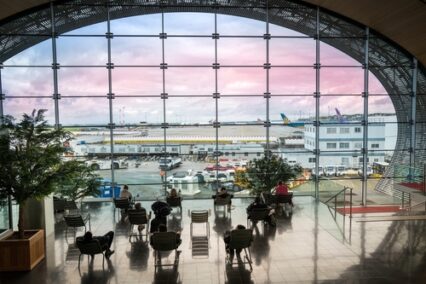Tourists seated in the departure lounge looking out at the airfield through a large oval window at Charles de Gaulle Airport.
