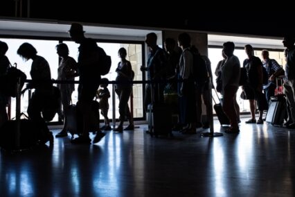 Silhouettes of passengers standing in line inside Aeropuerto de Sevilla, España, with luggage and carry-on bags, waiting for their boarding call.