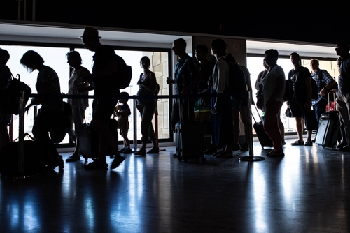 Silhouettes of passengers standing in line inside Aeropuerto de Sevilla, España, with luggage and carry-on bags, waiting for their boarding call.
