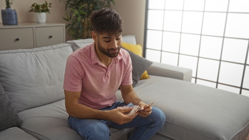 Young Hispanic man with a beard, sitting on a sofa in a living room, counting Mexican pesos, creating a relaxed and homely atmosphere indoors.