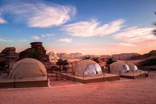 Three futuristic camping tents sit in the wadi rum desert of Jordan, a popular travel destination during sunset with dramatic blue sky and swirling cloud pattern.