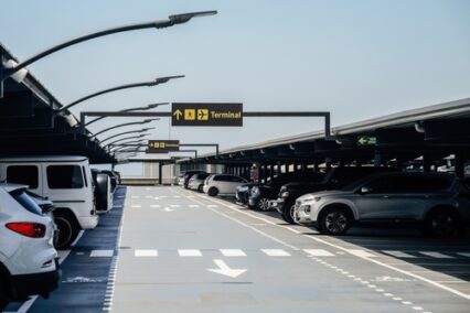 Modern airport parking lot with several cars parked under structured lighting and clear yellow signs directing passengers towards the terminal area