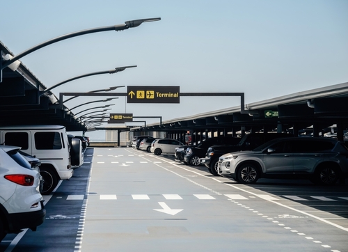 Modern airport parking lot with several cars parked under structured lighting and clear yellow signs directing passengers towards the terminal area