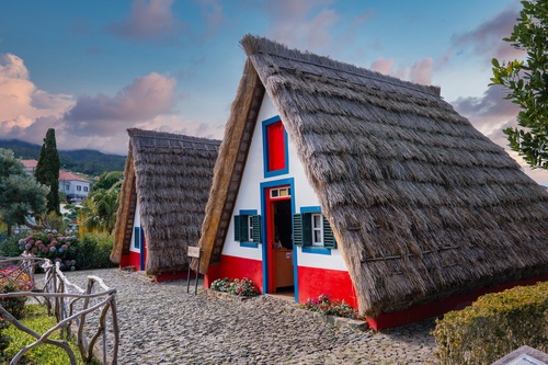 Two traditional A frame thatched houses with white walls and red bases stand by a cobblestone path in Santana, Madeira, Portugal