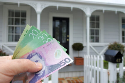 Man's hand holding New Zealand dollar currency bills against a traditional villa house in Auckland, New Zealand.
