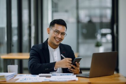 Happy businessman working on smartphone with accounting documents at the table in office.