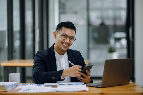 Happy businessman working on smartphone with accounting documents at the table in office.