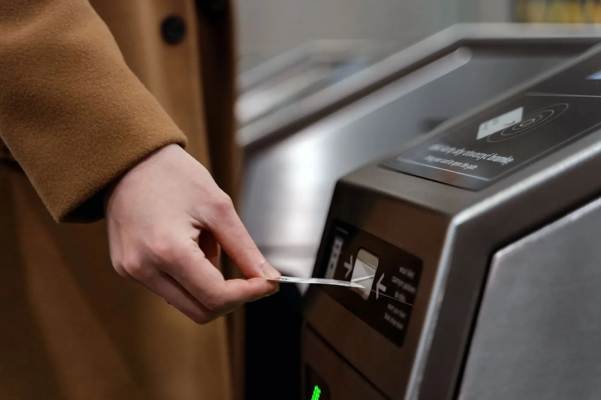 Hand in a tan wool coat inserting a paper ticket into a stainless steel metro turnstile