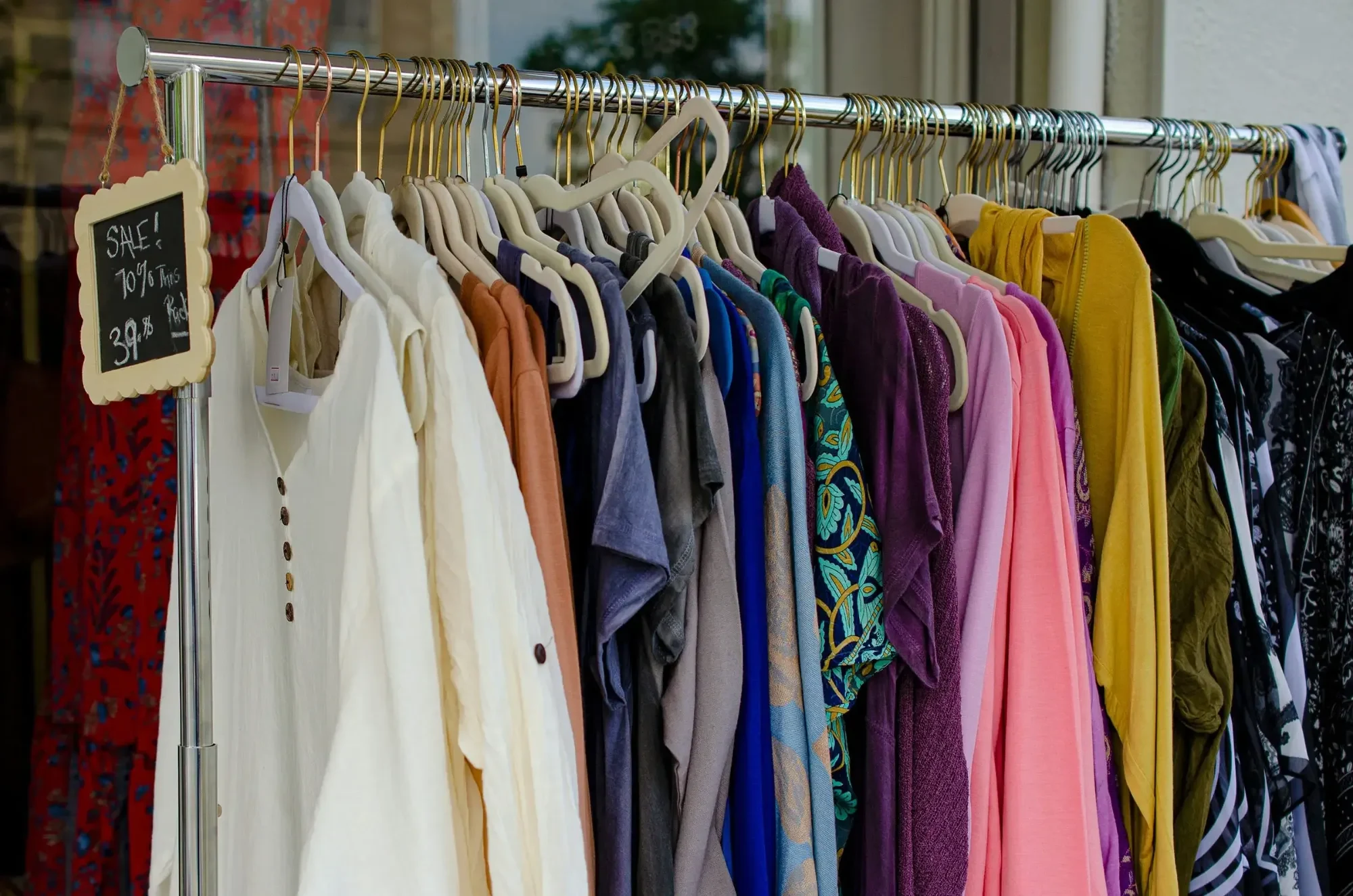 Rack of colorful secondhand clothes on hangers outside a shop, with a chalkboard sale sign.
